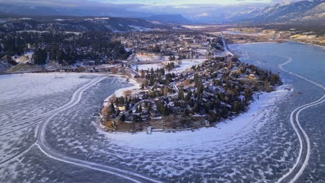 Aerial-drone-view-of-Windermere-Lake-frozen-over-passing-overhead-of-the-city-of-Windermere-with-ice-skaters-and-manmade-rinks-made-on-the-ice-below
