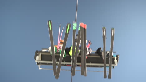 Looking-up-at-ski-lift,-family-of-colourful-skiers-passing-overhead-at-winter-sun-resort