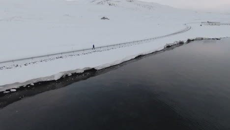 Orbit-view,-girl-walking-on-winding-snowy-road-by-calm-oceanshore,-Iceland
