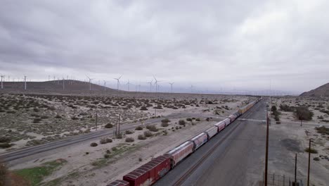 Aerial-Drone-Footage-of-Cargo-Train-in-Palm-Springs-Desert-with-Wind-Farms-in-the-Background,-slow-moving-pan