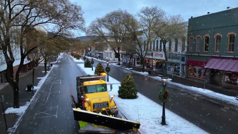 Snow-plow-driving-on-Wellsboro,-PA-main-street-lined-with-Christmas-decorations-during-active-snow-storm-in-December,-Beautiful-lit-Christmas-trees-and-stores-in-American-town