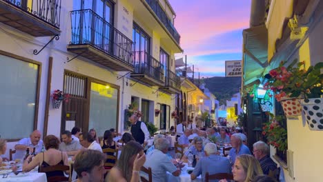 People-sitting-and-having-dinner-outside-in-a-restaurant-with-a-beautiful-pink-sunset-sky-in-Benahavis-Spain,-romantic-street-in-the-evening-during-summer,-4K-shot