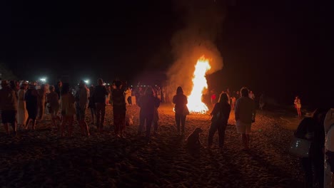 Big-crowd-at-the-traditional-bonfire-festival-at-the-beach-at-the-San-Juan-celebration-in-Marbella-Spain,-friends-and-family-enjoying-a-fun-party-during-summer,-burning-fire-and-hot-flames,-4K-shot