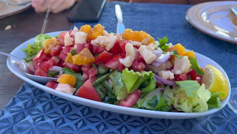 Squeezing-lemons-onto-a-delicious-and-healthy-salad-with-feta-cheese,-tomatoes,-lettuce,-onion-and-oranges,-tasty-lunch,-4K-shot