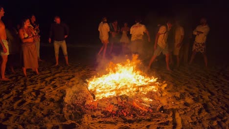 People-jumping-over-a-traditional-bonfire-at-the-San-Juan-festival-at-the-beach-in-Marbella-Spain,-friends-and-family-enjoying-a-fun-party,-jumping-over-a-burning-fire-and-hot-flames-at-night,-4K-shot