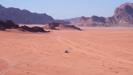 White-4WD-truck-following-tracks-across-red-sandy-vast-and-remote-desert-in-Wadi-Rum,-Jordan,-with-mountains-in-the-distance