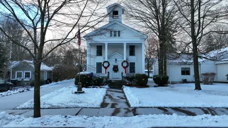 Slow-cinematic-aerial-dolly-forward-towards-old-white-church-building-while-snow-falls-in-small-town-in-America
