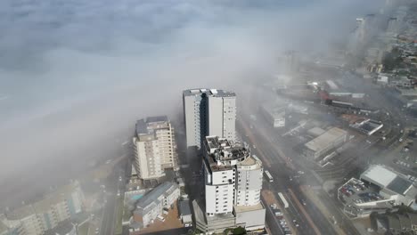 Dynamic-aerial-view-of-three-tall-buildings-in-a-busy-coastal-town-with-a-thick-layer-of-fog-covering-the-ocean-begind