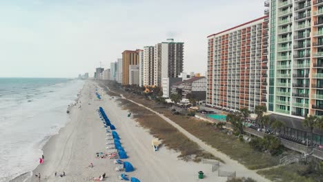-sandy-beach-lined-with-blue-umbrellas-stretching-out-towards-the-horizon-with-people-playing-in-the-waves-and-several-high-rise-buildings-in-the-background