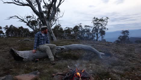 A-bushman-sits-around-a-camp-fire-on-a-fallen-down-snow-gum-up-in-the-mountains