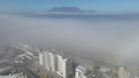 Aerial-footage-of-apartment-blocks-on-the-ocean-side-with-thick-fog-and-table-mountain-on-the-far-background