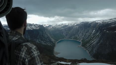Male-backpacker-on-mountaintop-enjoying-breathtaking-sea-view-with-snow-on-peak-during-cloudy-day-in-Norway