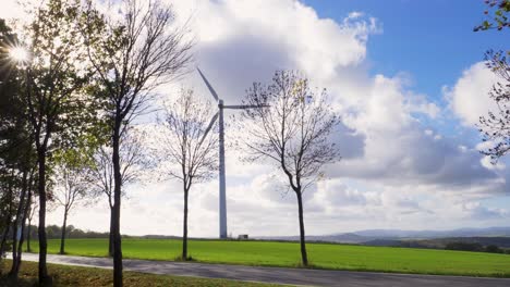wind-turbine-standing-still-on-hill-blue-sky-with-white-clouds,-sun-breaks-through-trees