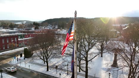 Slow-aerial-orbit-around-bright-American-flag-waving-on-sunny-winter-day-with-snow-in-small-town,-Wellsboro-PA