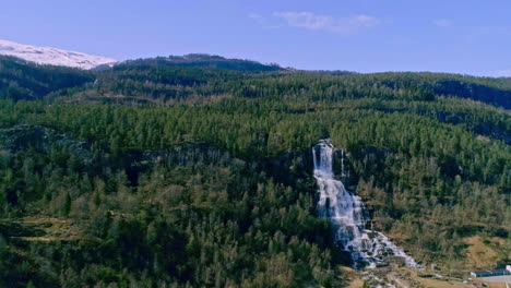 Aerial-backwards-shot-of-natural-waterfall-flowing-down-green-forest-mountains-durign-sunny-day-in-Norway---Snow-on-peak-in-background