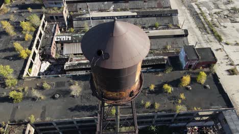 Rusty-water-tower-with-open-rooftop-hatch-in-abandoned-building-complex,-aerial-ascend-view