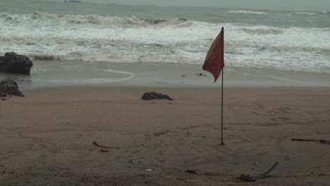 Rote-Fahne-Auf-Einem-Dünnen-Stock,-Der-Im-Winter-An-Einem-Strand-Mit-Wellen-Im-Hintergrund-Im-Sand-Steckt