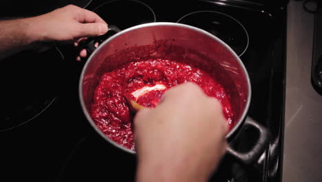 Male-hands-stirring-juicy-cranberries-in-saucepan-boiling-sauce-on-kitchen-stove