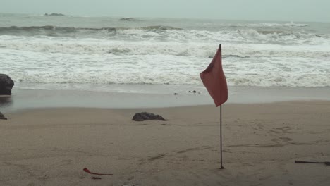Bandera-Roja-De-Advertencia-De-Playa-Y-Salvavidas-Atrapada-En-La-Arena-Con-Olas-Rompiendo-En-La-Orilla-En-El-Fondo