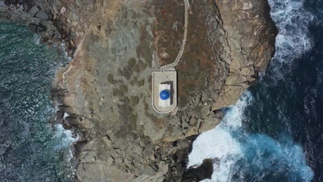 An-aerial-birds-eye-view-of-a-small-church-in-a-greek-island,-between-the-cliffs-and-the-sea