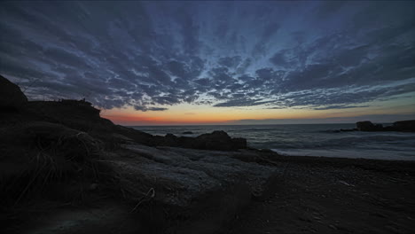 Low-Angle-Timelapse-Am-Strand-Mit-Sonnenuntergang-Am-Horizont-über-Dem-Meer-Mit-Vorbeifliegenden-Wolken