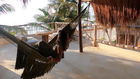 Drone-shot-of-a-girl-swinging-on-a-hammock-on-a-rooftop-with-a-beautiful-beach-view-in-Tulum