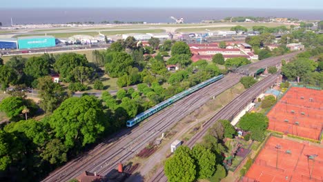 Dolly-in-aerial-view-of-the-Buenos-Aires-train,-sustainable-city-transportation,-airplane-landing-strip-in-the-background,-sunny-day