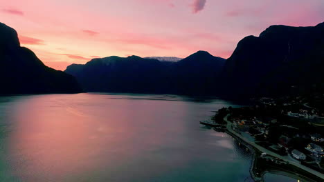 Stunning-aerial-shot-of-golden-hour-pink-light-that-reflects-in-the-lake-and-black-silhouettes-of-the-mountains-in-buhe-background-of-a-norwegian-village
