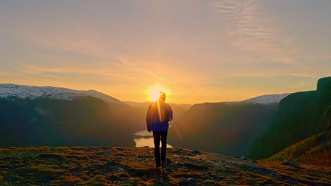 Hiker-In-Hoodie-Jacket-Walking-On-Top-Of-A-Mountain-Overlooking-Fjord-At-Sunrise
