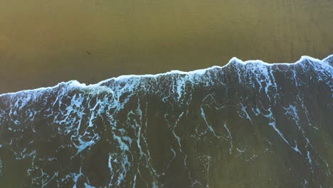 A-drone-captures-a-dramatic-view-of-the-ocean's-energy-as-waves-crash-onto-the-sandy-shore,-creating-frothy-white-foam