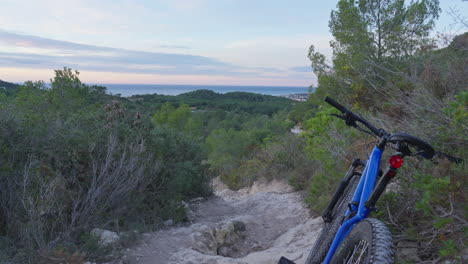 Mountain-bike-parked-on-dirt-track-leading-downhill-toward-stunning-early-morning-sky-and-sea-views-at-horizon