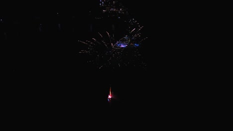 An-aerial-point-of-interest-rotation,-around-exploding-fireworks-during-the-night-from-the-sea,-by-a-pier