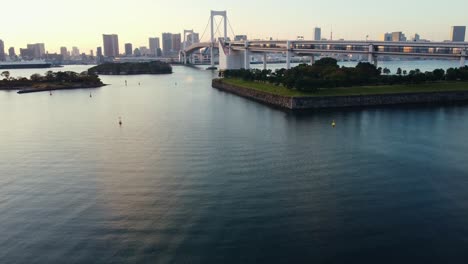 Aerial-Drone-Flying-Low-Over-Water-Revealing-Rainbow-Bridge-In-Odaiba-Tokyo-City-During-Sunset