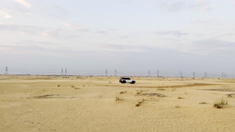 SUV-Blanco-Conduciendo-Rápido-Por-El-Desierto-Al-Atardecer-Con-Cables-Eléctricos-En-El-Fondo