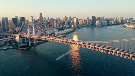 Aerial-Drone-Flying-Low-Over-Water-Towards-Rainbow-Suspension-Bridge-in-Odaiba-Tokyo-City-Japan-While-Boat-Sails-Under-The-Bridge-with-Tokyo-Tower-in-the-Background