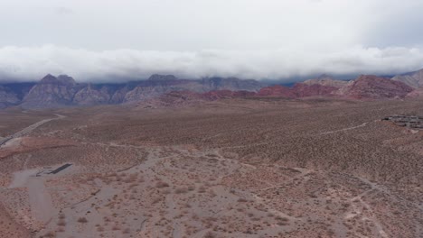 Wide-descending-aerial-shot-of-Red-Rock-Canyon-with-thick-clouds-over-the-mountains-in-Las-Vegas,-Nevada