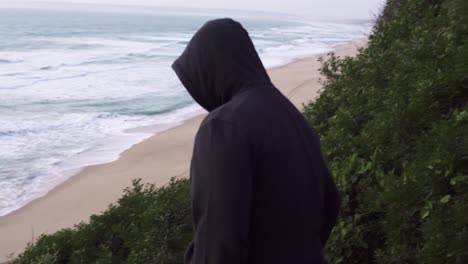 Hooded-person-dressed-in-black-going-down-a-cliff,-beach-with-waves-in-the-background-at-sunset