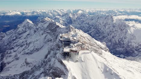 Dramatische-Luftaufnahme-Des-Gebäudes-Auf-Einem-Schneebedeckten-Gipfel-Mit-Den-Sich-Entfaltenden-Alpen-Im-Hintergrund