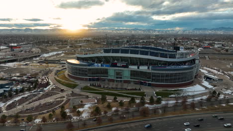 Drone-flying-away-from-iconic-Empower-field-at-Mile-High-Stadium-formerly-Broncos-Stadium-and-revealing-cars-driving-through-main-street-highway-and-Downtown-Denver-city-view-backdrop-during-sunset