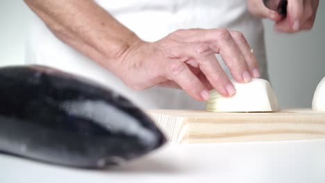 Senior-caucasian-person-cutting-onion-with-a-knife-on-a-wooden-cutting-board,-fish-in-defocused-foreground