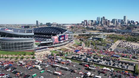 Cinematic-aerial-shot-of-iconic-Empower-field-at-Mile-High-Stadium-with-cars-parked-in-parking-spaces-and-main-street-highway-and-Downtown-Denver-city-view-backdrop,-Colorado