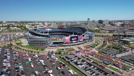 Cinematic-drone-shot-revealing-Empower-field-at-Mile-High-Stadium-exterior-with-cars-parked-in-parking-spaces,-main-street-highway-with-Downtown-Denver-city-view-backdrop,-Colorado