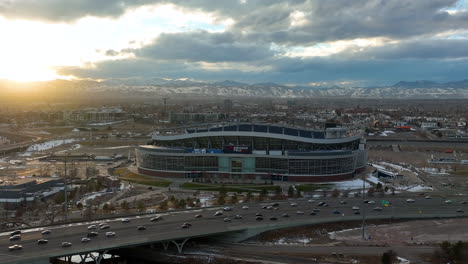 Cinematic-drone-shot-of-iconic-Empower-Field-at-Mile-High-formerly-known-as-Broncos-stadium-with-a-view-of-main-street-and-snow-capped-mountain-in-background,-Colorado