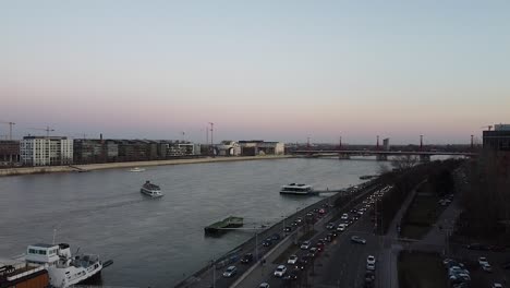 Luftaufnahme-Des-Verkehrs-Bei-Sonnenuntergang-Mit-Einem-Schiff,-Das-Auf-Der-Donau-In-Budapest-Treibt,-Mit-Der-Stadt-Im-Hintergrund