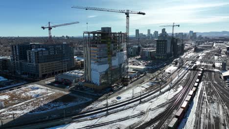 Drone-flying-close-to-multi-storey-building-under-construction-site-with-view-of-rail-track-covered-with-snow-in-and-downtown-denver-city-view-in-background,-Colorado