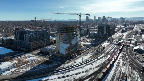 Cinematic-aerial-view-of-multi-story-building-construction-site-with-view-of-rail-tracks-partially-covered-with-snow-in-foreground-and-city-view-in-background