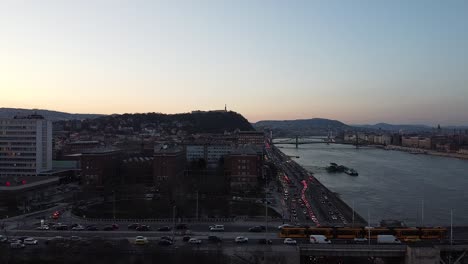 Luftaufnahme-Des-Verkehrs-Bei-Sonnenuntergang-Mit-Der-Donau-In-Budapest-Mit-Der-Stadt-Und-Den-Bergen-Im-Hintergrund