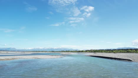Flying-low-upstream-over-braided-Waimakariri-River-in-New-Zealand---beautiful-turquoise-water,-blue-sky-and-mountains-on-low-horizon
