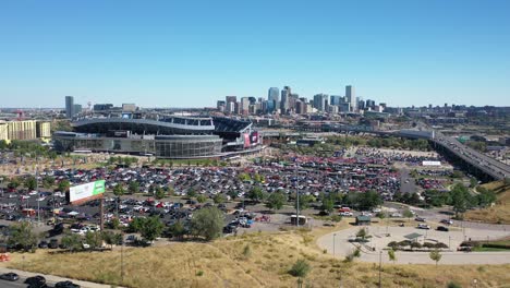 Cinematic-aerial-tracking-view-of-Empower-field-at-Mile-High-Stadium-with-lots-of-cars-parked-in-parking-spaces-and-main-street-highway-and-Downtown-Denver-city-view,-Colorado