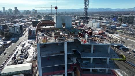 Drone-flying-around-multi-storey-buildings-under-construction-site-and-workers-working-on-roof-with-city-view-at-background,-Denver
